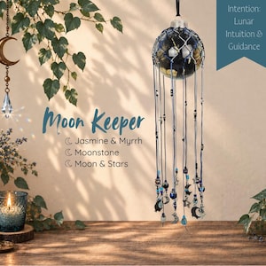 May include: A hanging Moon Keeper ornament featuring a dark glass sphere encased in blue netting with dangling blue cords, beads, and metal moon and star charms. Text on the image reads Moon Keeper, Jasmine & Myrrh, Moonstone, Moon & Stars, and Intention: Lunar Intuition & Guidance.