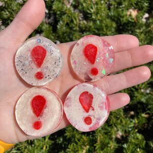 Puede incluir: Cuatro posavasos redondos de resina transparente con un diseño de signo de exclamación rojo. Dos posavasos tienen purpurina plateada y los otros dos tienen purpurina iridiscente. Los posavasos se sostienen en una mano.