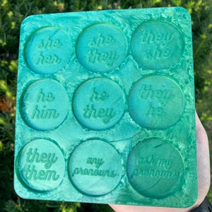 May include: A green tray with nine round compartments, each with a different pronoun phrase embossed in white. The phrases are "she her", "she they", "they she", "he him", "he they", "they he", "they them", "any pronouns", and "ask my pronouns".