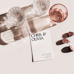 May include: A styled photograph featuring a wedding invitation with the names "Chris & Olivia" printed on it. Two crystal glasses, a pink glass, and sliced figs are arranged on a light surface, with shadows cast by sunlight.