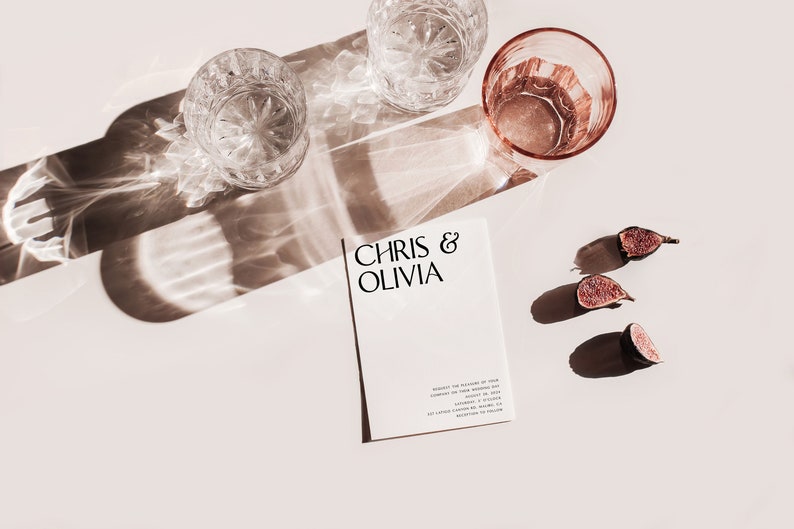 May include: A styled photograph featuring a wedding invitation with the names "Chris & Olivia" printed on it. Two crystal glasses, a pink glass, and sliced figs are arranged on a light surface, with shadows cast by sunlight.