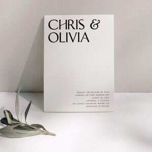May include: A minimalist wedding invitation card with the names "CHRIS & OLIVIA" in bold black font. The card includes the wedding date, time, and location details in a smaller font. A sprig of greenery rests near the card.