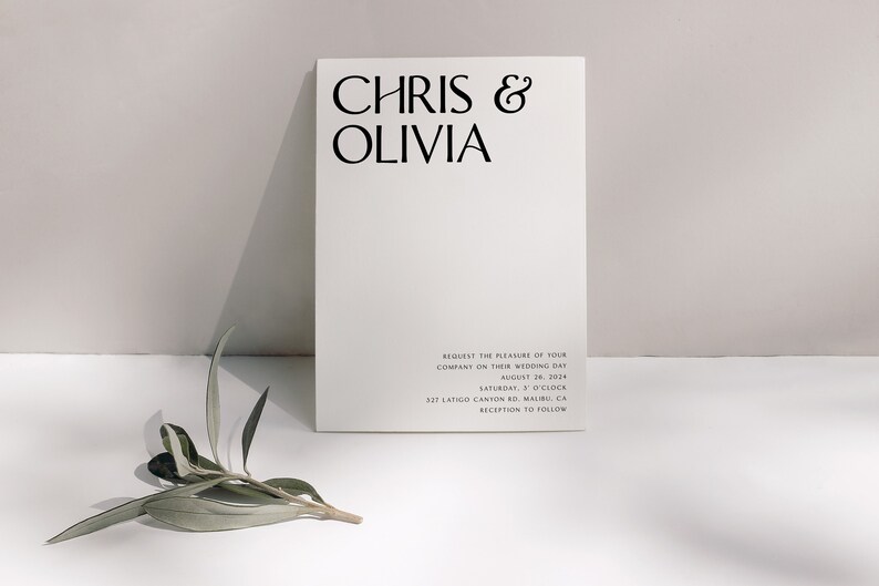 May include: A minimalist wedding invitation card with the names "CHRIS & OLIVIA" in bold black font. The card includes the wedding date, time, and location details in a smaller font. A sprig of greenery rests near the card.