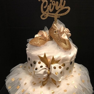 May include: A white diaper cake decorated with gold glitter and polka dots. The cake is topped with a pair of gold glitter baby shoes and a gold glitter sign that says "It's a Girl".