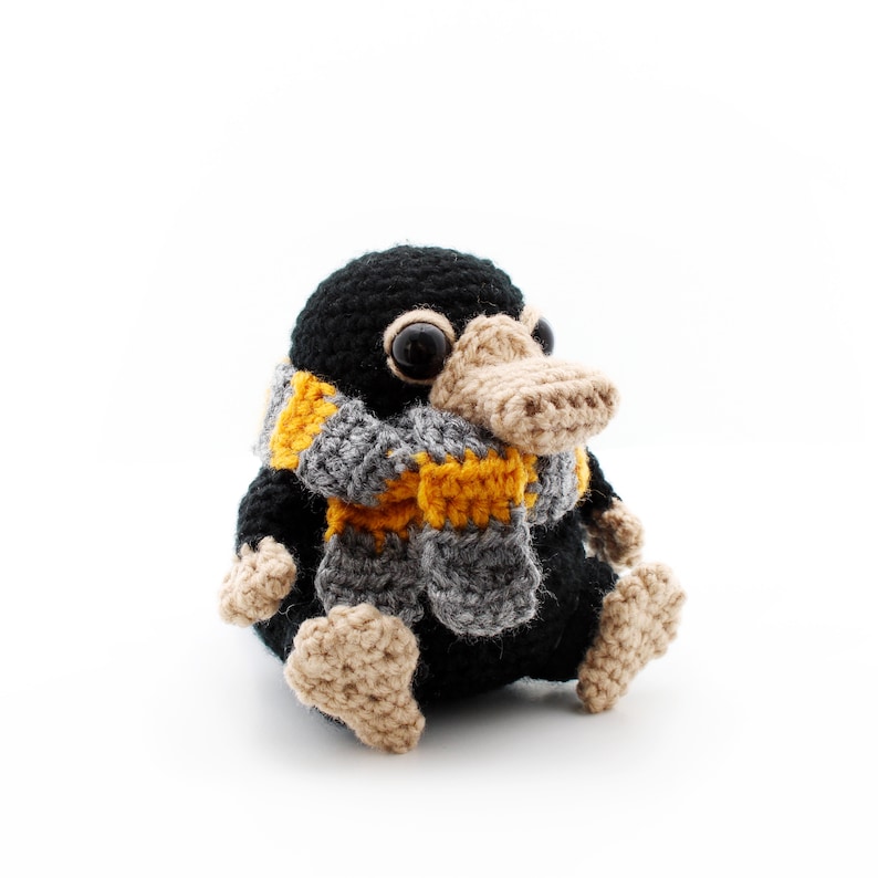 May include: A black crocheted Niffler plush toy wearing a yellow and grey scarf. The Niffler has a long snout and large, black eyes.