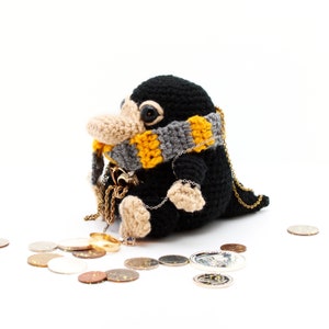 May include: A black and brown crocheted Niffler plush toy wearing a yellow and grey scarf. The Niffler is sitting on a white surface with gold coins and a gold ring scattered around it.