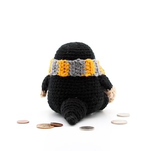 May include: A black crocheted mole with a yellow and grey striped scarf sitting on a white surface with several coins scattered around it.