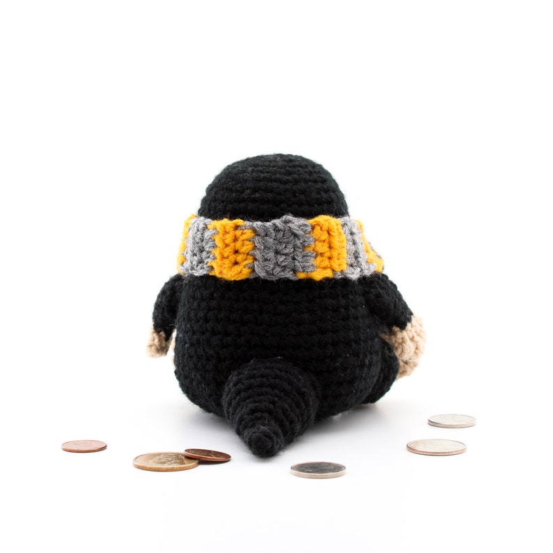 May include: A black crocheted mole with a yellow and grey striped scarf sitting on a white surface with several coins scattered around it.