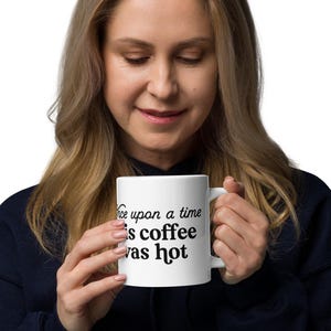 May include: White ceramic mug with the text "Once upon a time is coffee was hot" in black. The mug is held by a person wearing a dark blue sweatshirt.