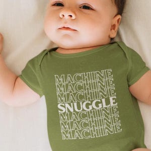 May include: An olive green baby bodysuit with the words "MACHINE SNUGGLE MACHINE MACHINE MACHINE" printed in white. The baby is lying on a white surface.