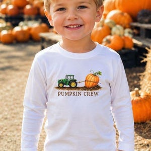 May include: A white long-sleeve shirt featuring a graphic of a green tractor pulling an orange pumpkin with the text PUMPKIN CREW printed in brown letters below the illustration. The shirt has a crew neckline and is made of a solid white fabric.