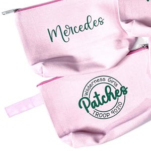 May include: Pink canvas zippered pouches. One pouch has the name "Mercedes" in green script. Another pouch features a circular design with the words "Wilderness Girls Patches TROOP 90210" in green.