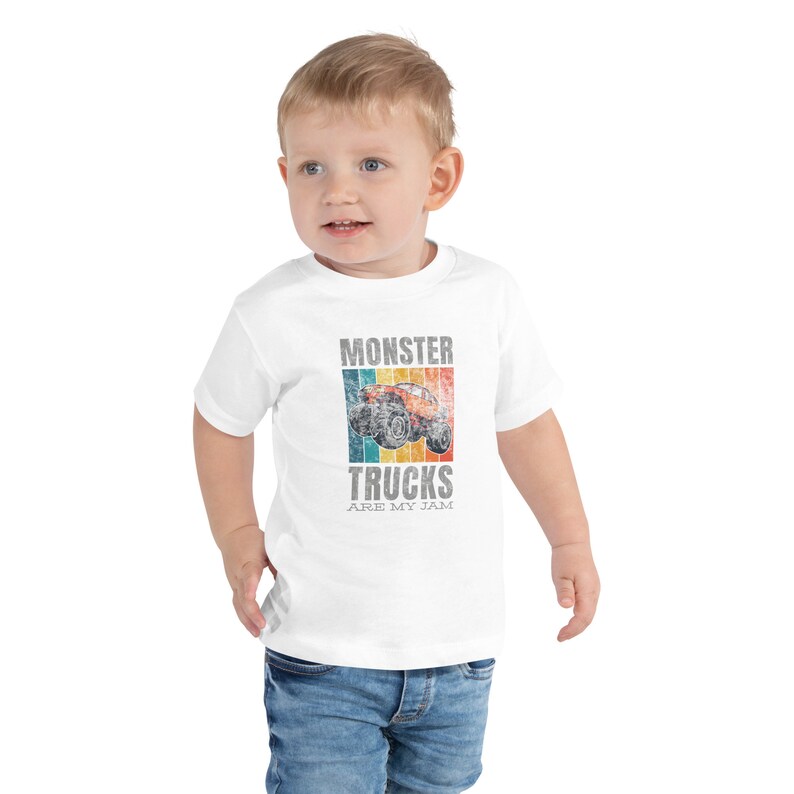 May include: A white toddler t-shirt with a retro-style monster truck graphic and the words "MONSTER TRUCKS ARE MY JAM". The design is in shades of orange, blue, and yellow. The child is wearing blue jeans.