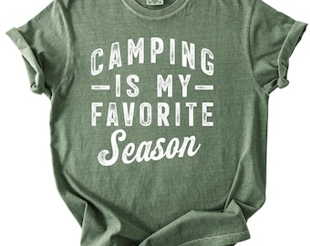 May include: Olive green t-shirt with rolled sleeves. The shirt features the white text "CAMPING IS MY FAVORITE SEASON" in a bold, vintage-style font. The shirt is made of a soft material and is suitable for casual wear.