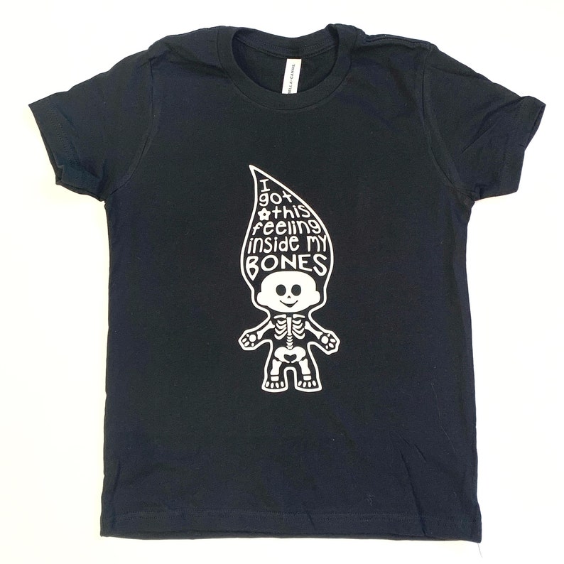 May include: A black short-sleeved t-shirt featuring a white graphic of a troll doll with a skeleton ribcage and pelvis. The troll's hair is shaped like a flame and contains the text: I GOT THIS feeling inside MY BONES, with a small flower icon.