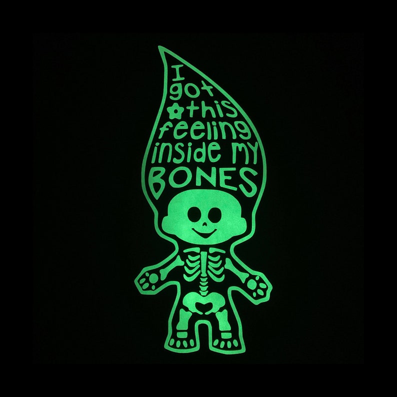 May include: A glow-in-the-dark graphic of a troll doll with a skeleton ribcage and pelvis design. The tall, pointed hair features the text I got this feeling inside my BONES with a small flower icon. The entire illustration is bright neon green.