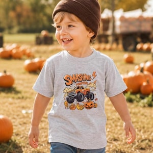 May include: A light gray short-sleeved t-shirt featuring a graphic print of an orange monster truck driving over pumpkins. The text SMASHING PUMPKINS is printed in orange above the truck. The design includes orange lightning bolts and pumpkin illustrations.