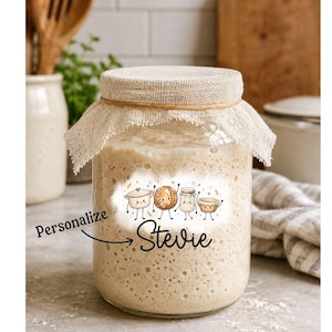 May include: A clear glass jar filled with bubbly sourdough starter, topped with a beige burlap fabric cover secured by a thin twine. The front features a graphic of four cartoon kitchen items and the text Personalize and Stevie in black.