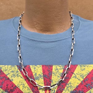 Pode incluir: Um colar de corrente de prata com um design de corrente de caixa. A corrente está em um manequim que está vestindo uma camiseta azul com um design vermelho, amarelo e branco.