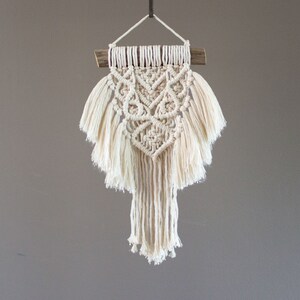 Peut inclure: Une suspension murale en macramé blanche avec un motif géométrique et des franges. La suspension est fixée à un petit bâtonnet en bois.