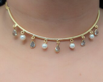 May include: A gold-toned choker necklace featuring a curved metal bar adorned with alternating dangling white spherical pearls and clear, pear-shaped faceted crystals suspended from small jump rings along the entire length of the collar.