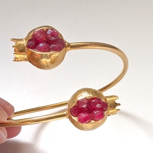 May include: A gold bangle bracelet with two pomegranate-shaped charms. Each charm is filled with red gemstone beads.