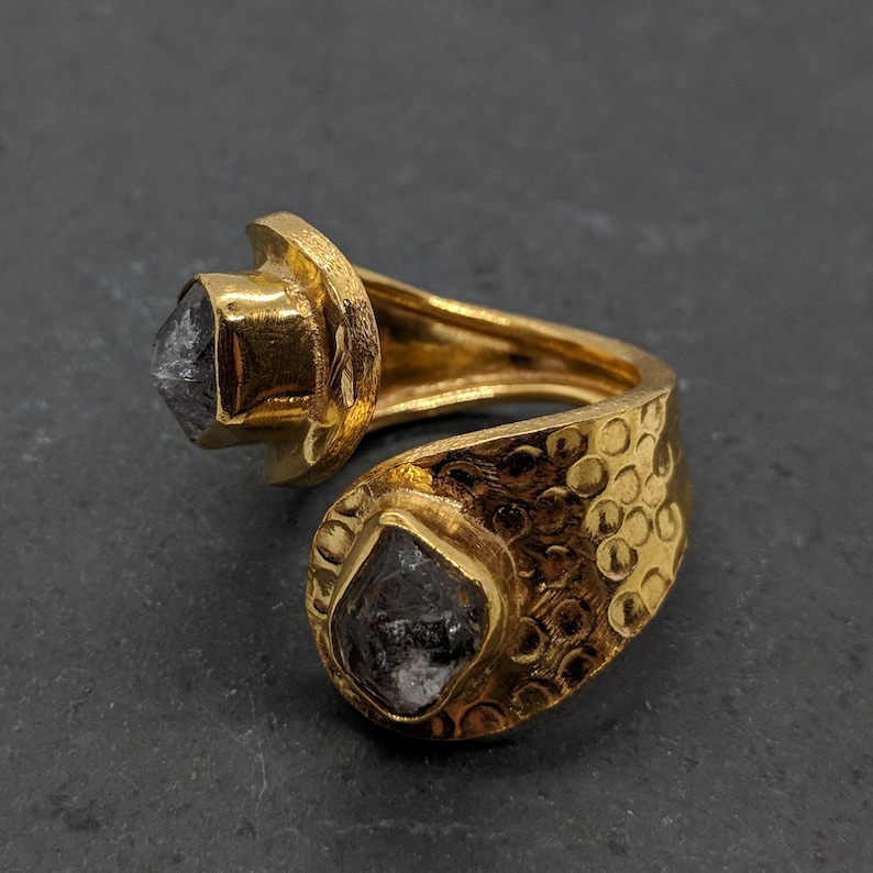 May include: A gold-toned open ring featuring a hammered texture on the band and two bezel-set, raw, dark gray, multifaceted stones at each end. The metal surface has a reflective finish, and the stones are irregular in shape and size.