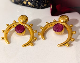 May include: A pair of gold-colored earrings with a crescent moon design. Each earring features a round, deep red gemstone set in the center of the crescent. Small gold beads adorn the curve of the crescent, and a decorative gold element sits at the top.