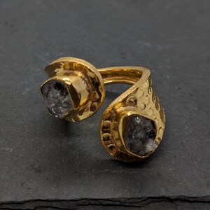 May include: An open-style gold ring featuring a hammered texture on the band and two prong-set, raw, dark gray stones at each end. The metal has a polished, reflective finish, and the stones have an irregular, natural, and faceted appearance.