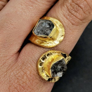May include: A gold-toned open ring featuring two hammered, crescent-shaped settings, each holding a raw, dark gray, translucent gemstone. The metal surface has a textured, uneven finish, and the two ends of the ring do not meet, creating a gap.