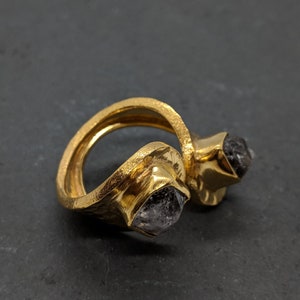May include: A gold-toned open ring featuring a textured, hammered metal band. The ring is set with two raw, dark gray, multifaceted stones, each held in a square-shaped bezel setting at the open ends of the band, creating a distinct bypass design.
