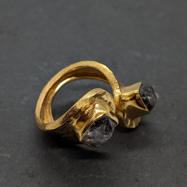May include: A gold-toned open ring featuring a textured, hammered metal band. The ring is set with two raw, dark gray, multifaceted stones, each held in a square-shaped bezel setting at the open ends of the band, creating a distinct bypass design.