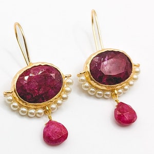 Può includere: Un paio di orecchini d'oro con una grande pietra di rubino di forma ovale incastonata in una cornice d'oro testurizzata. Gli orecchini sono decorati con una fila di piccole perle bianche e una piccola pietra di rubino che pende dalla parte inferiore.