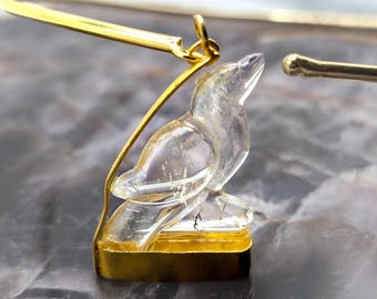 May include: A clear crystal bird pendant with gold accents. The bird is perched on a gold base and is suspended from a gold chain.