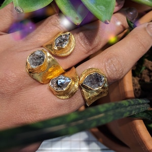 May include: Four gold-toned rings with textured, uneven bands, each featuring a single, raw, translucent grey crystal set in a prominent bezel. The rings are worn on fingers, showcasing the metallic finish and the natural, irregular shape of the stones.