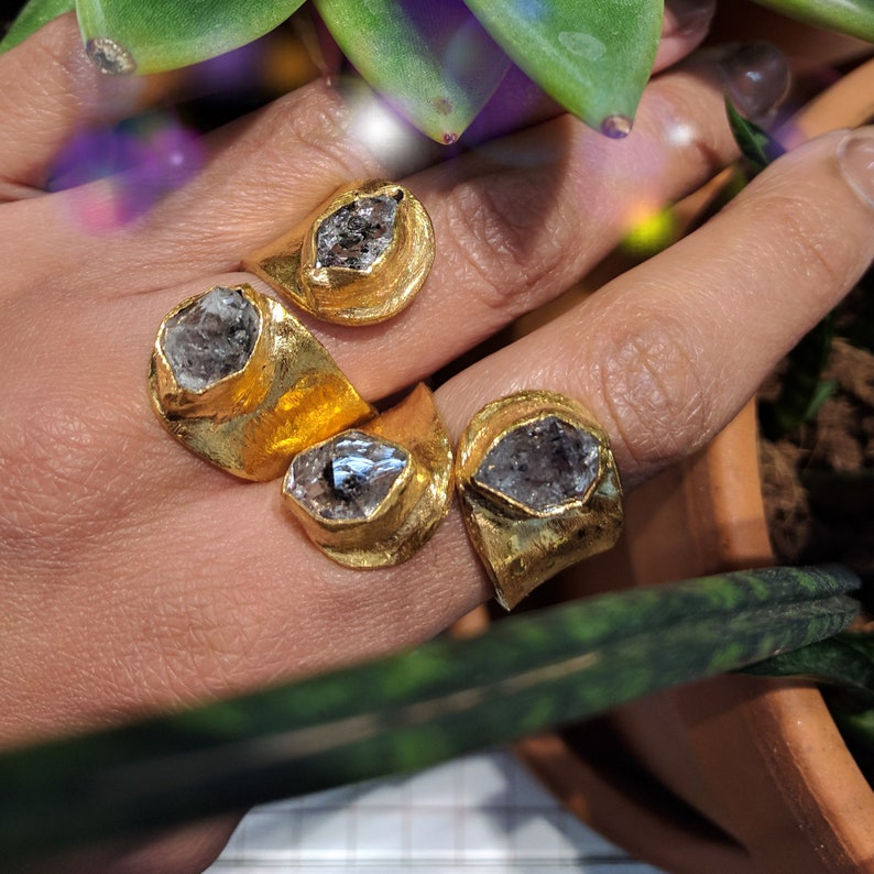 May include: Four gold-toned rings with textured, uneven bands, each featuring a single, raw, translucent grey crystal set in a prominent bezel. The rings are worn on fingers, showcasing the metallic finish and the natural, irregular shape of the stones.