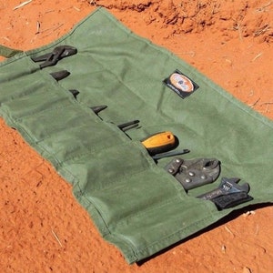 May include: A green canvas tool roll containing a selection of tools, including pliers, screwdrivers, and wrenches. The roll is unfurled and laid out on a red dirt surface.