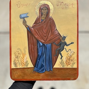May include: A hand-painted icon depicting a woman in a red robe and blue dress holding a hammer and a small black devil. The icon is on a wooden panel with a gold background and the text "S. ZINPT" and "M. 22.17" in the top corners.