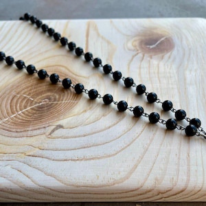 May include: A black beaded necklace with a silver cross pendant. The necklace is on a light wood surface.