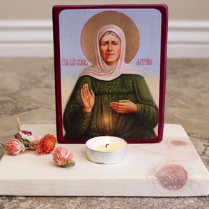 May include: A wooden icon stand with a small candle and dried flowers. The icon is of a woman in a green dress and white head covering. The text on the icon reads "Святая Екатерйна" and "Аттирья".