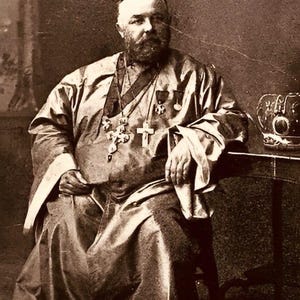 May include: Sepia-toned portrait of a man in religious attire, seated and looking at the viewer. He wears a long robe, multiple necklaces with crosses, and medals. A small table with a decorative object is to his right.