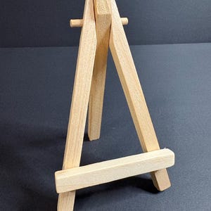 May include: A small, natural wood easel with a three-legged design. The easel is made of light-colored wood and is designed to hold small canvases or artwork. The easel is freestanding and has a simple, minimalist aesthetic.