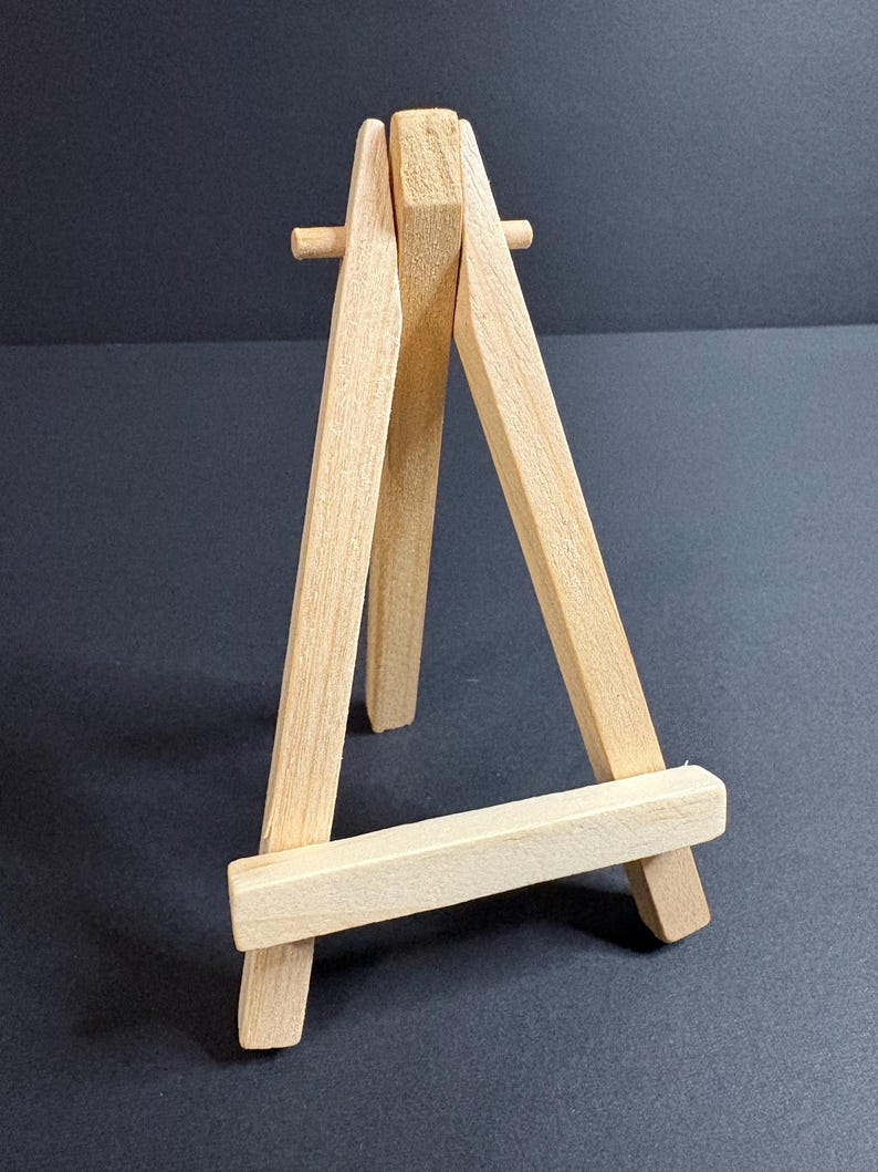 May include: A small, natural wood easel with a three-legged design. The easel is made of light-colored wood and is designed to hold small canvases or artwork. The easel is freestanding and has a simple, minimalist aesthetic.