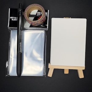 May include: A collection of art supplies on a black surface. Includes a black pen, a roll of decorative tape, a pack of clear sleeves, and a small white canvas on a wooden easel. The tape has a black and white checkered pattern.