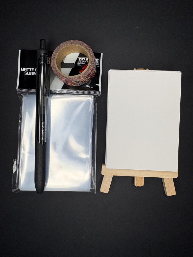 May include: A collection of art supplies on a black surface. Includes a black pen, a roll of decorative tape, a pack of clear sleeves, and a small white canvas on a wooden easel. The tape has a black and white checkered pattern.