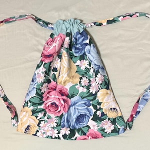 May include: A floral drawstring backpack featuring a pattern of pink, blue, and yellow roses with green leaves and white flowers. The bag has a light blue top and matching floral straps.