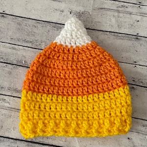 Può includere: Un cappello a forma di mais caramellato all'uncinetto con la parte superiore bianca, la parte centrale arancione e la parte inferiore gialla.