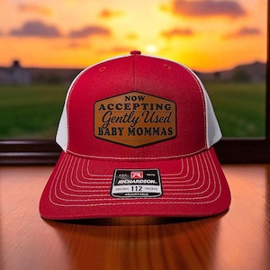 May include: Red and white trucker hat with the text "NOW ACCEPTING Gently Used BABY MOMMAS" on a brown patch. The hat has a curved brim and an adjustable strap. The background shows a sunset.