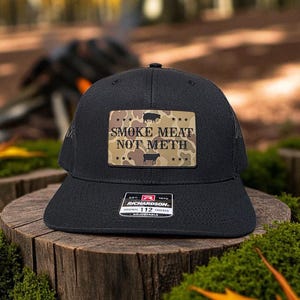May include: Black trucker hat with a mesh back and a flat brim. The front features a rectangular patch with the text "SMOKE MEAT NOT METH" above a camouflage pattern with pig silhouettes. The hat has a Richardson 112 tag.