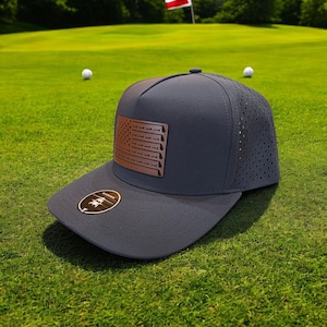May include: A navy blue baseball cap with a brown leather American flag patch on the front. The flag has a unique design with golf club cutouts. The cap has a mesh back and a curved brim.